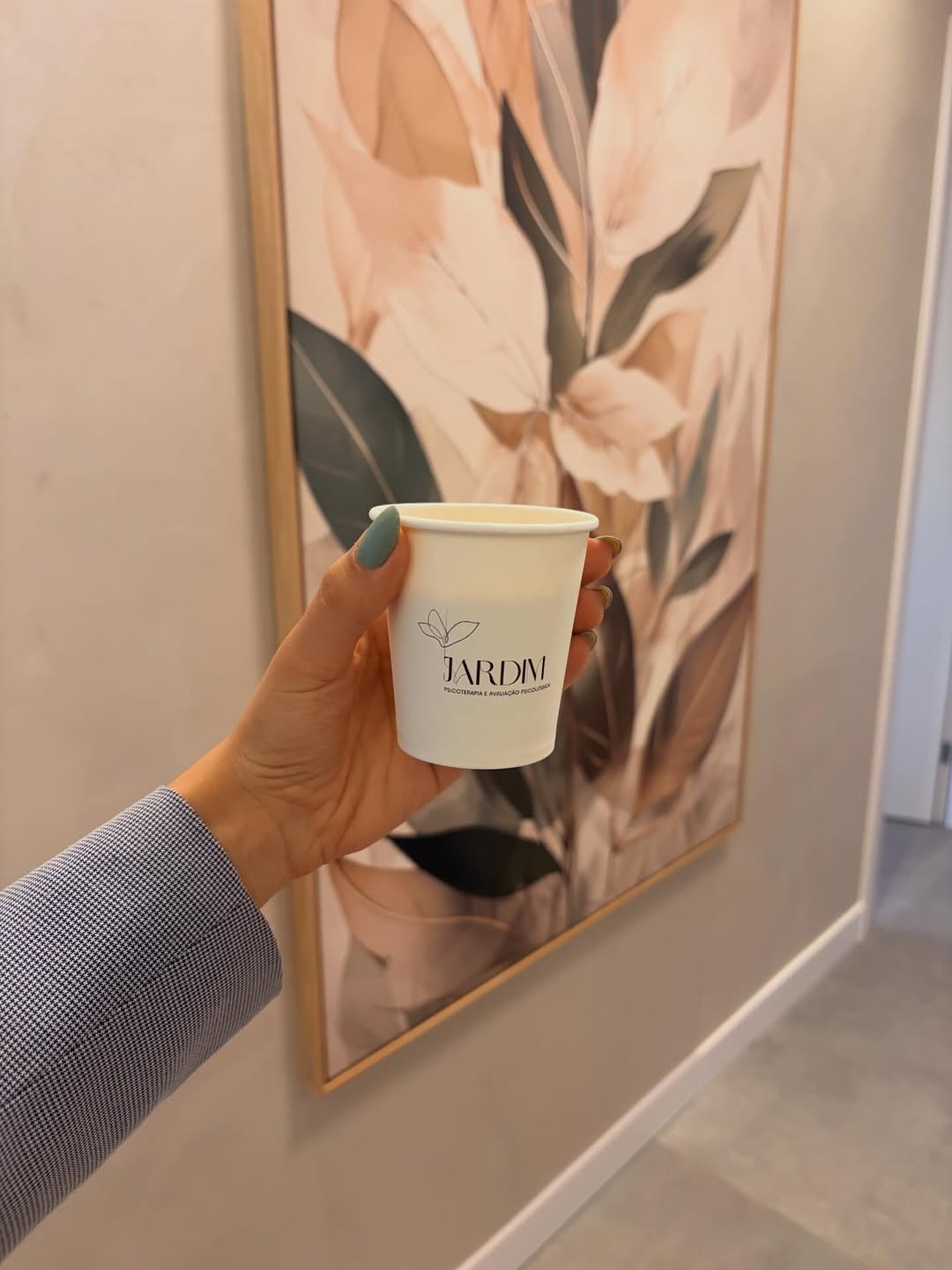 Copo com o logotipo da Jardim na mão, diante do quadro de folhas da recepção.