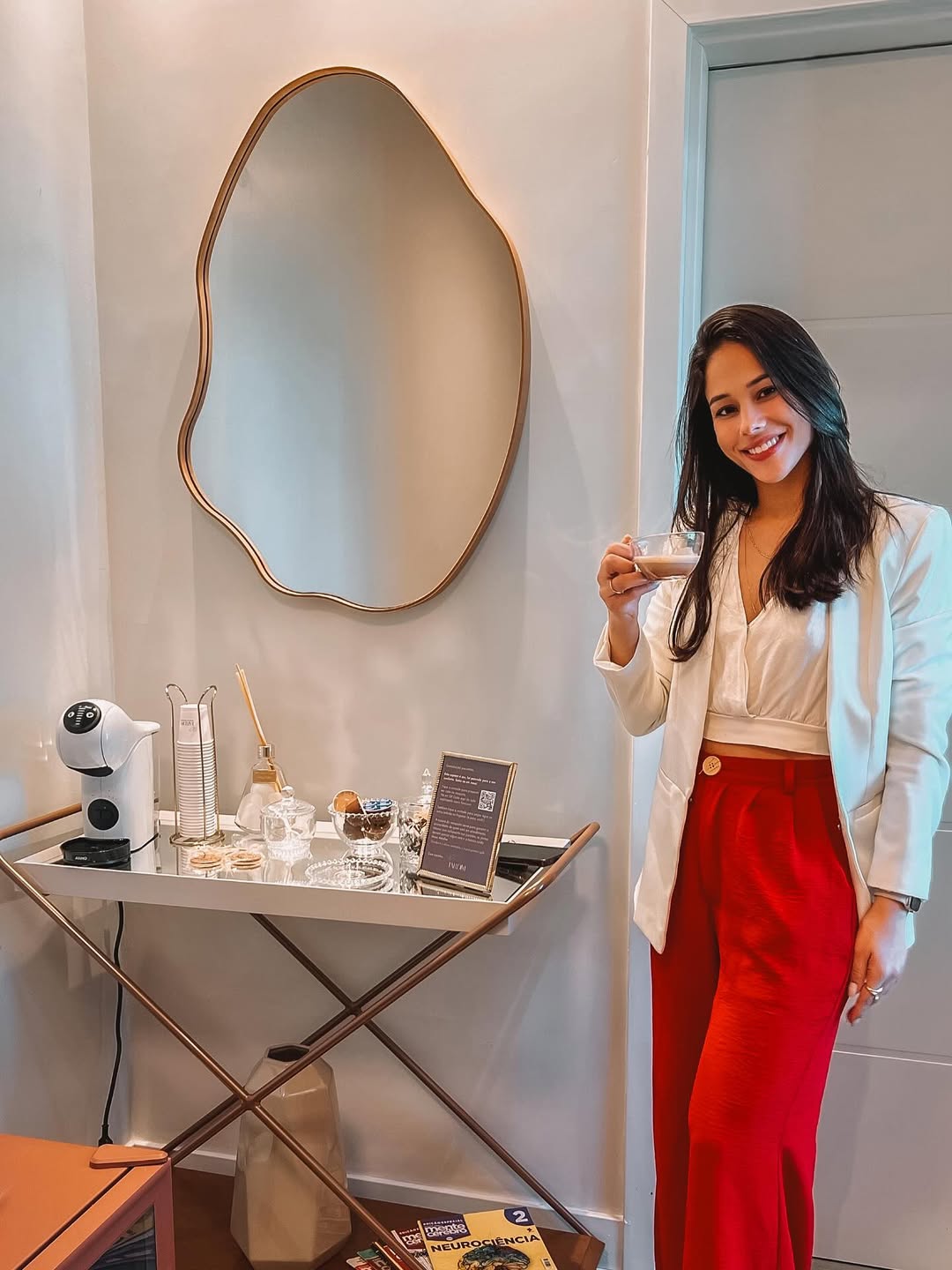 Genara Mello na recepção da Clínica Jardim, de blazer branco, com uma xícara de café na mão, ao lado do espelho dourado e do carrinho de café.